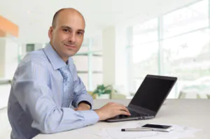 A man in a shirt looking at the camera with one hand on a laptop in front of him